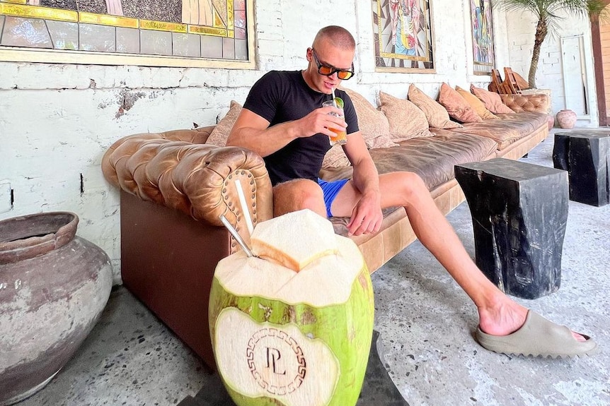 A man sips a drink sitting on a couch, with a coconut sitting on a table in front