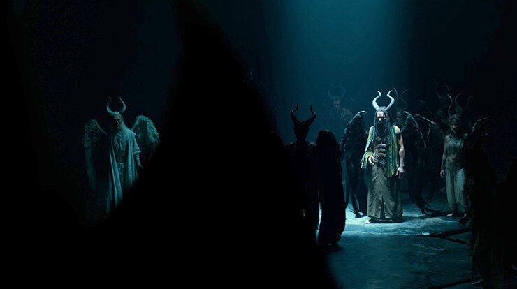 Chiwetel Ejiofor wears large horns and wings and stands under spotlight in a dark cavernous space near other similar creatures.