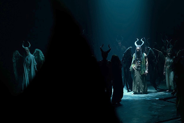 Chiwetel Ejiofor wears large horns and wings and stands under spotlight in a dark cavernous space near other similar creatures.
