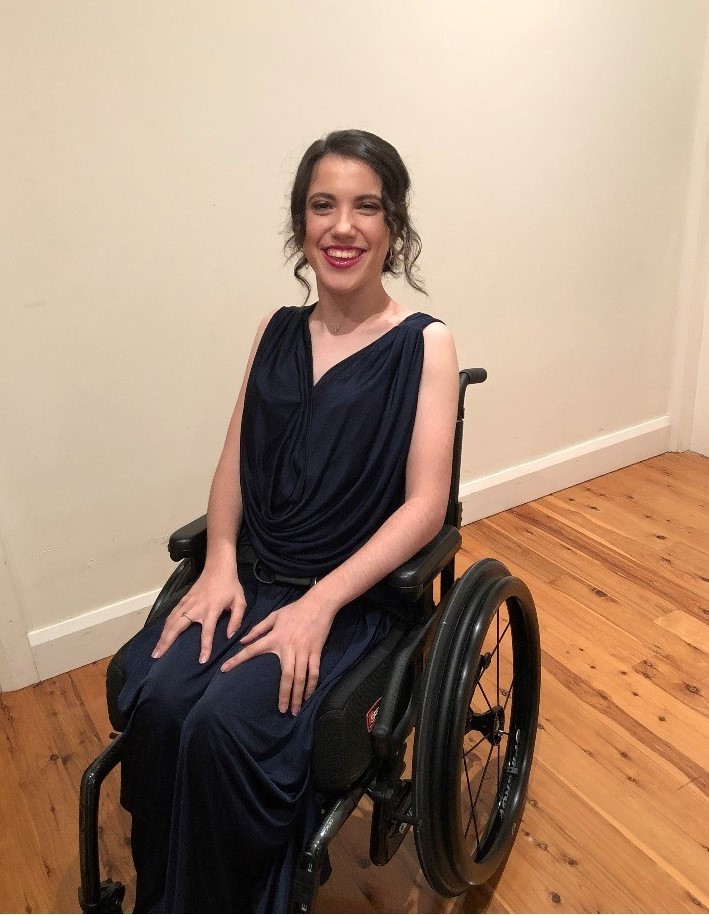 A young woman, wearing a dark-coloured jumpsuit, suits smiling in a wheelchair
