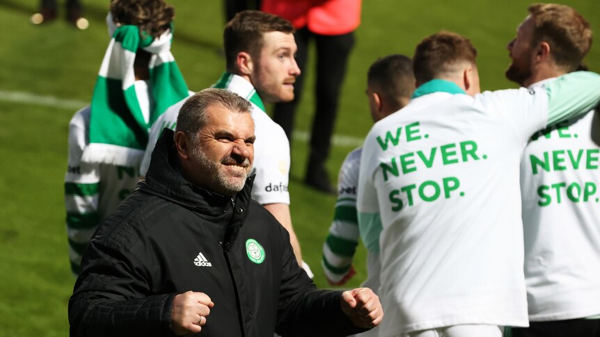 Celtic manager Ange Postecoglou grins and pumps his fists in celebration as players hug in the background after winning a title.