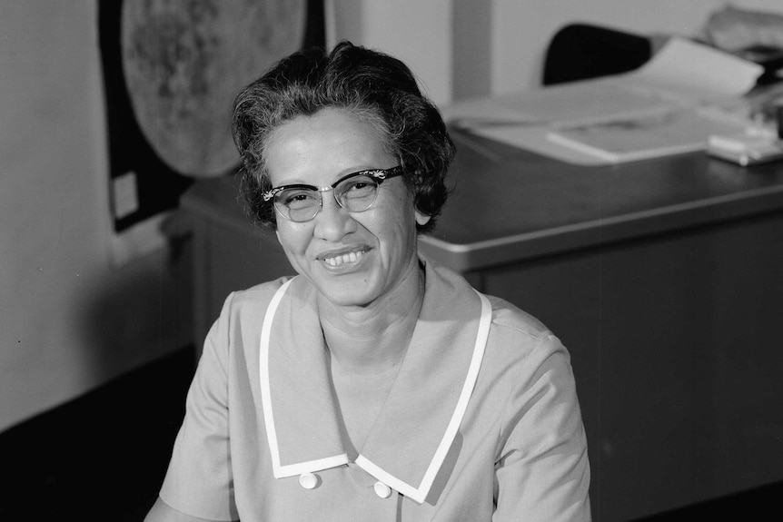 A black and white photo of NASA research mathematician Katherine Johnson at her desk smiling in 1966.