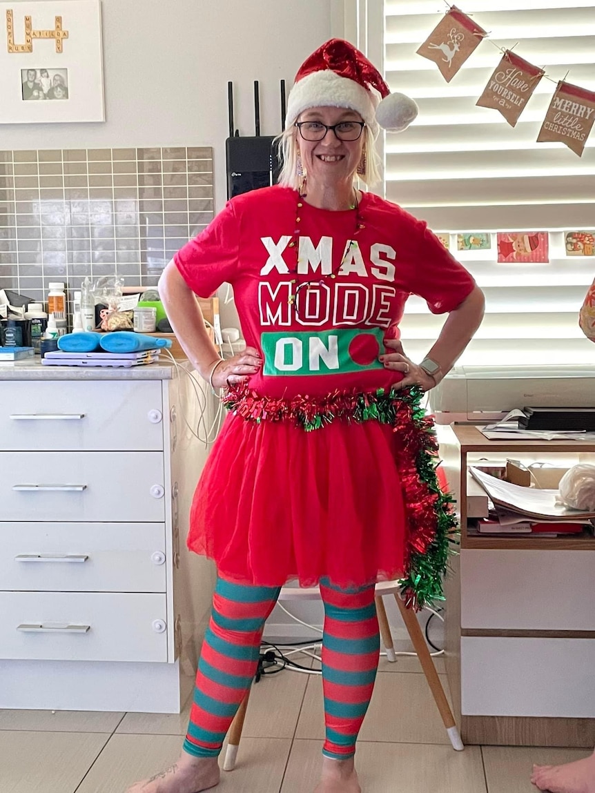 A woman, wearing a red Christmas t-shirt and a Santa hat, stands smiling in her kitchen.