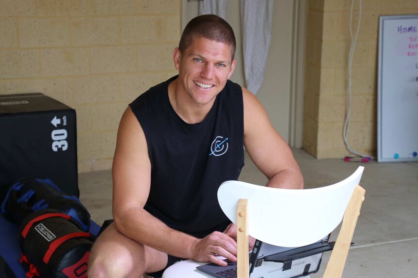 A personal trainer wearing a black tank top sits smiling for a photo in a garage with a laptop on a chair in front of him.