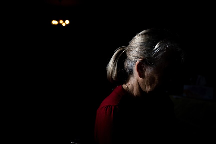 A woman sits in a dimly lit room. Her face is covered in shadow, her head is lit from the back.