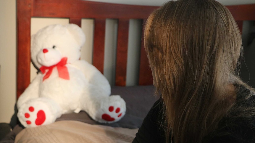 A woman with her face obscured looks at a white teddy bear on a bed.