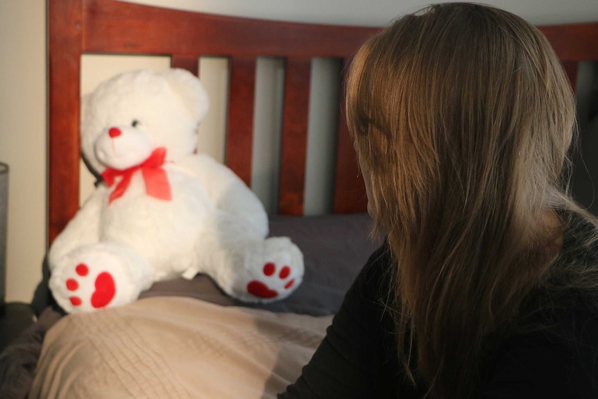 A woman with her face obscured looks at a white teddy bear on a bed.