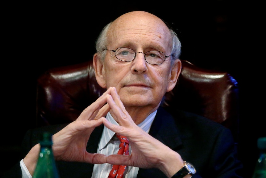 An old man in glasses presses his fingers together in front of him on a desk, looking interested.