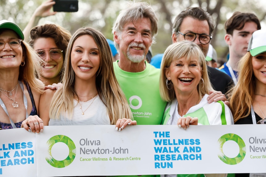 Olivia Newton-John stands alongside her husband and daughter, all smiling, in a crowd of people holding up a banner