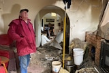 Brad wears a red waterproof jacket and jeans as he surveys the huge damage inside his house.