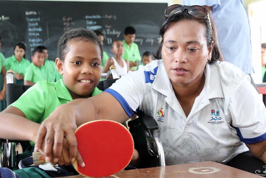 Harvi crouches down, showing a young girl in a wheelchair how to use the table tennis paddle.