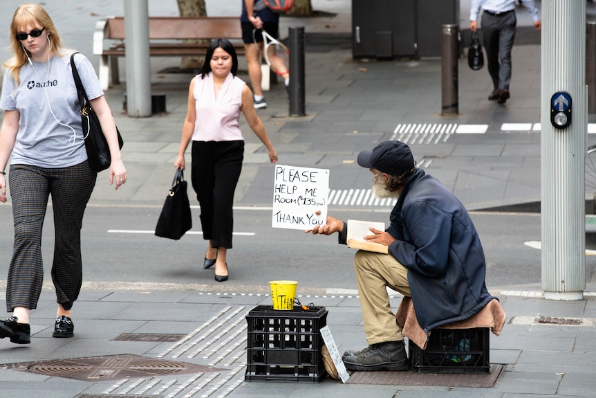 A homeless man on a crate holding a sign and wearing a hat, jacket
