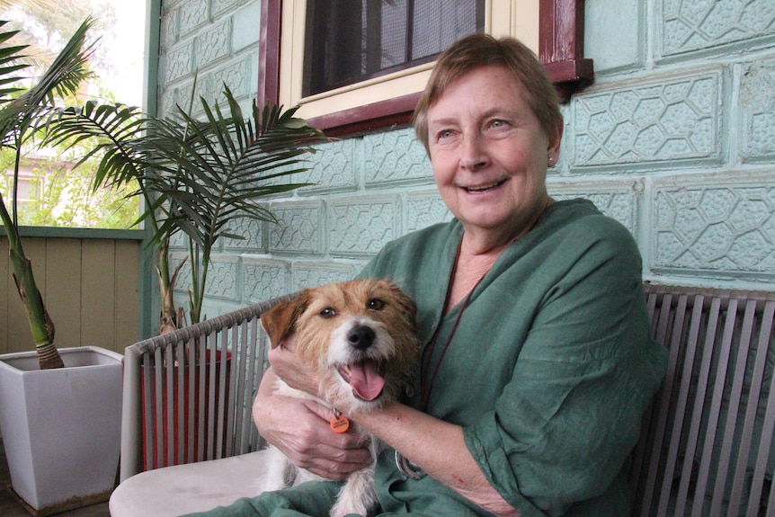 A woman sits outside her house holding her wire haired Jack Russell terrier with plants in the background.