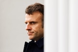 Emmanuel Macron stands behind a door frowning in the distance.