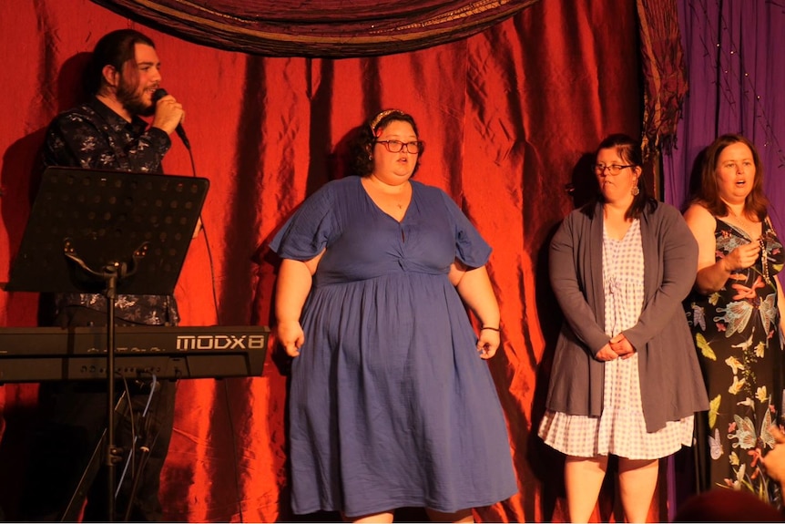 A man and three women stand on stage in a line in front of a red curtain, he has a microphone