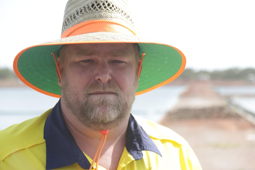 A man in a bright yellow work shirt and hat looks seriously at the camera.