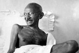 Mahatma Ghandi sits on a bed laughing.