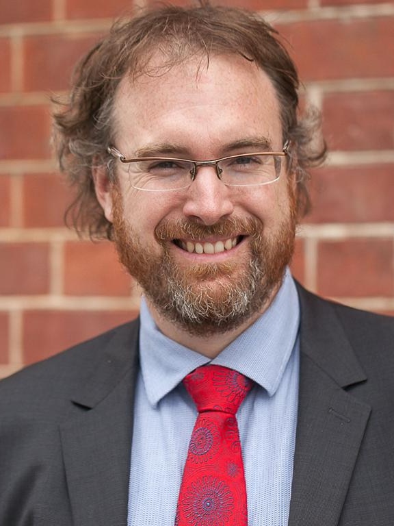 A man with glasses, a blue shirt and red tie and a grey suit jacket standing in front of a brick wall