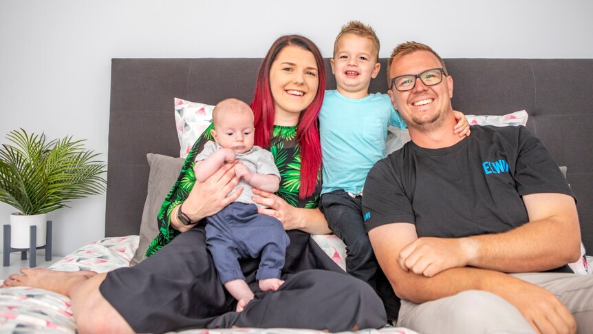 A family of four on a bed smiling: The mother has red hair and is holding a baby, the father is wearing glasses