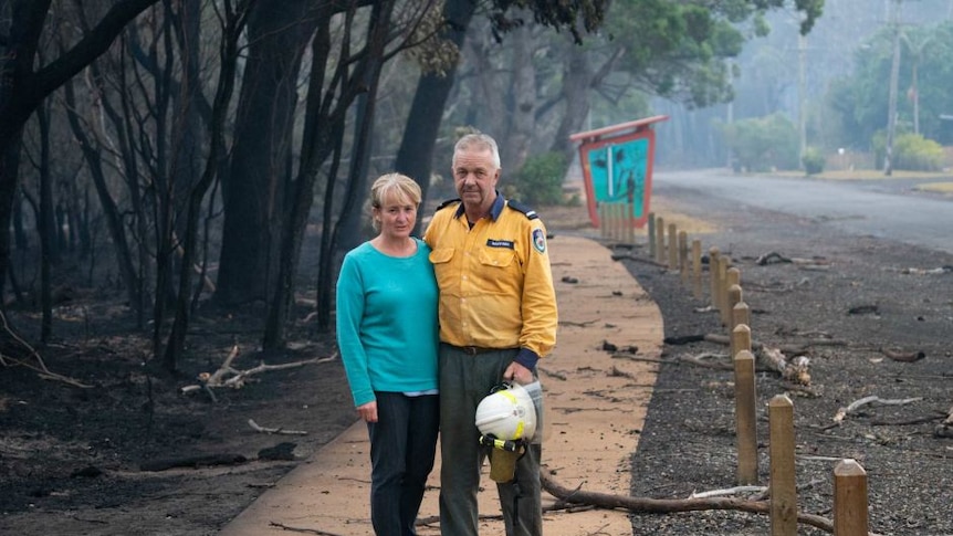 A man in a firefighters uniform and a woman pose with arms around one another on a footpath next to burnt out forest.