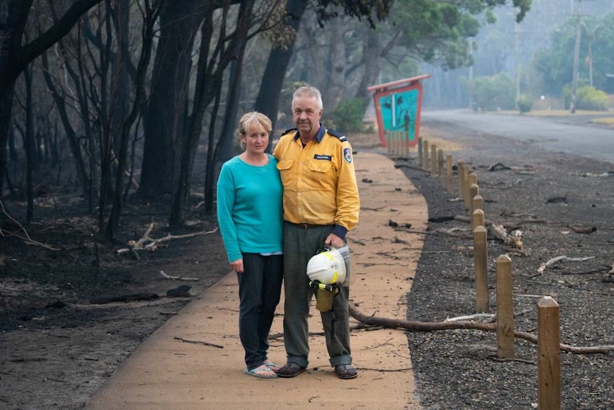 A man in a firefighters uniform and a woman pose with arms around one another on a footpath next to burnt out forest.