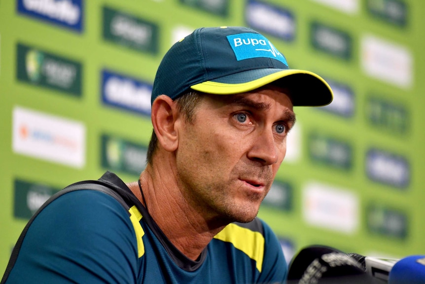 Justin Langer, in his Australian training gear, purses his lips as he answers a question at a press conference.