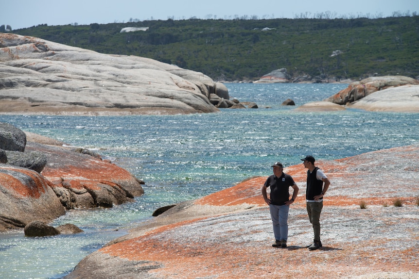 Two people stand on the rocks by a glistening blue ocean.