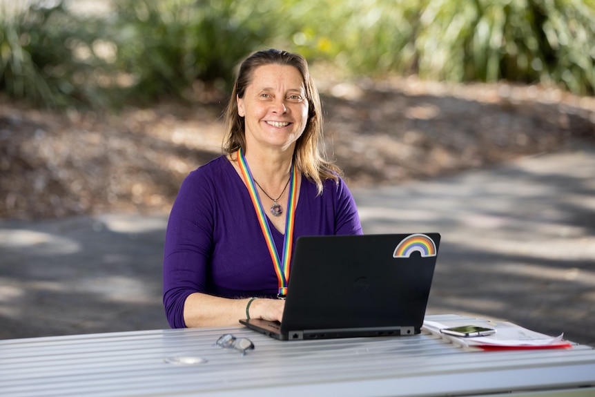 A smiling, dark-haired woman sitting with her laptop in a pleasant outdoor area. 