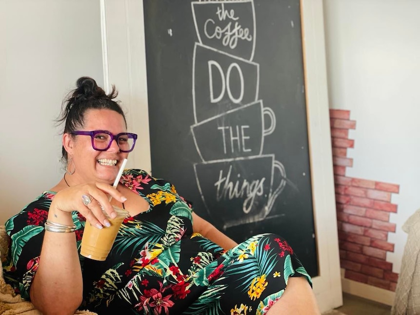 A woman with dark hair and purple glasses sitting in a cafe with an iced coffee.