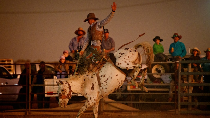 A man riding a bucking horse in an outback rodeo arena, at dusk.