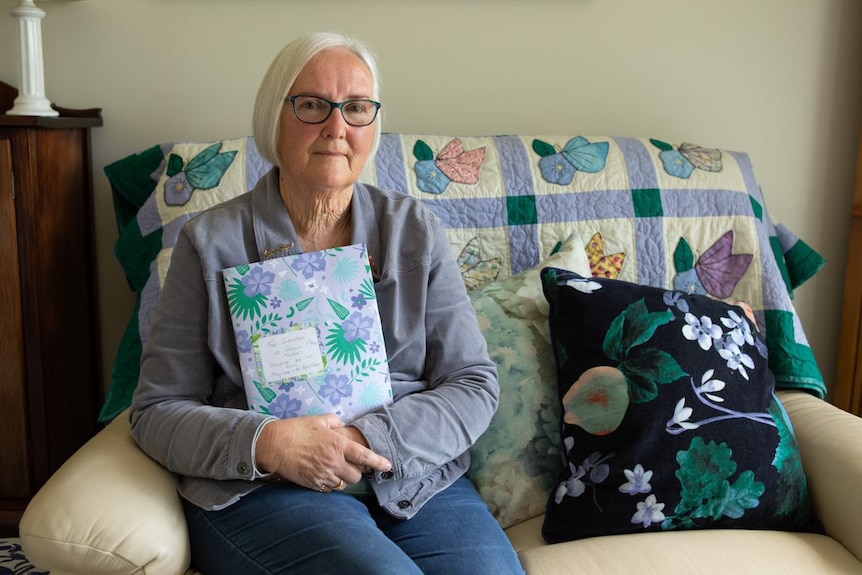 A woman with white hair and glasses holds a diary.