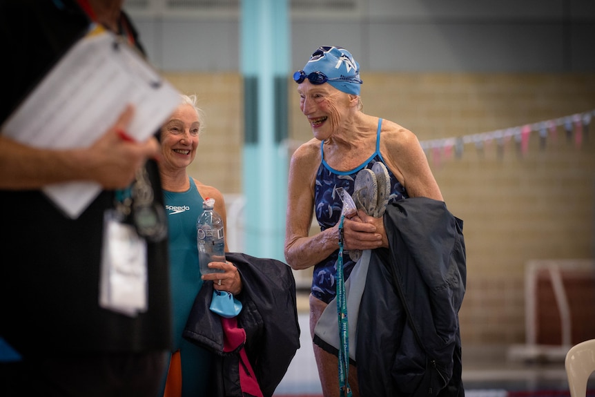 An elderly woman in a swim suit holds her towel and laughs