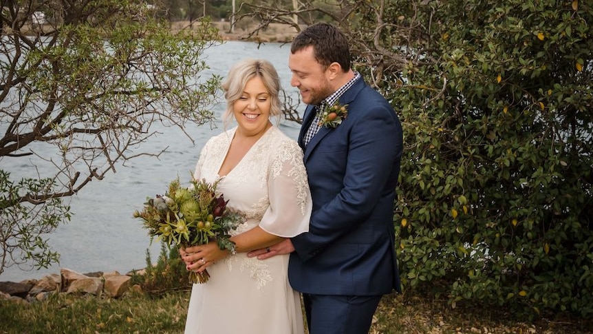 A bride and groom smile while standing on the edge of a lake.