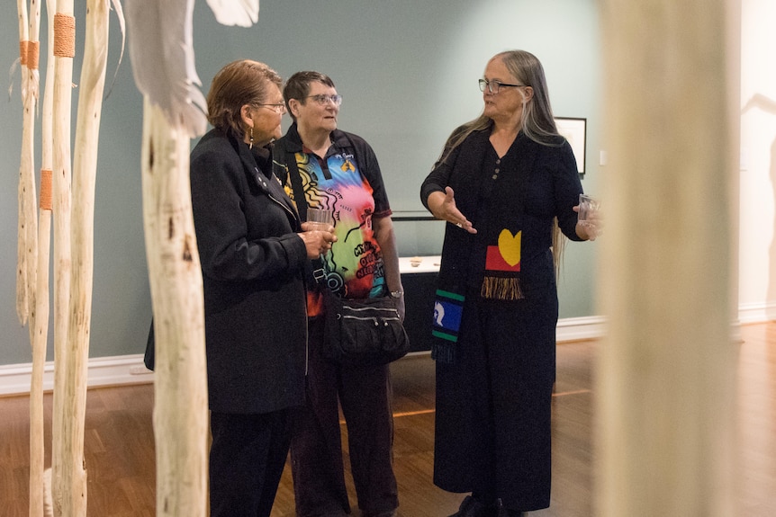 Three indigenous artists chat at an exhibition opening, surrounded by cultural objects and artworks