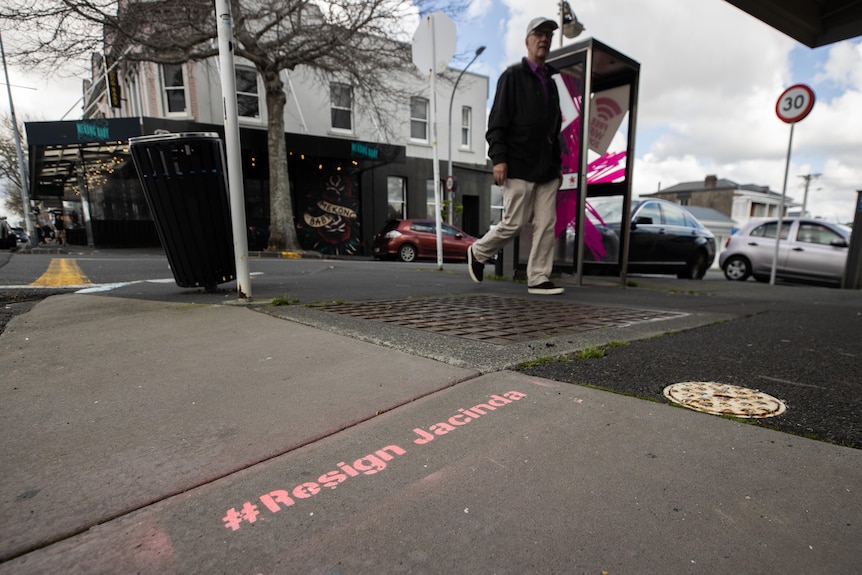A stencil of the words #ResignJacinda is painted in pink on a cement footpath with a man walking past. 