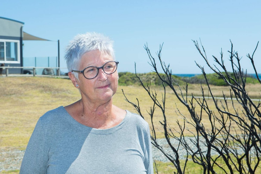 A woman with shirt grey hair standing next to a burnt bush near the beach