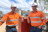 Two men in hi vis shirt and hard hats stand in front of structures at a mining site.