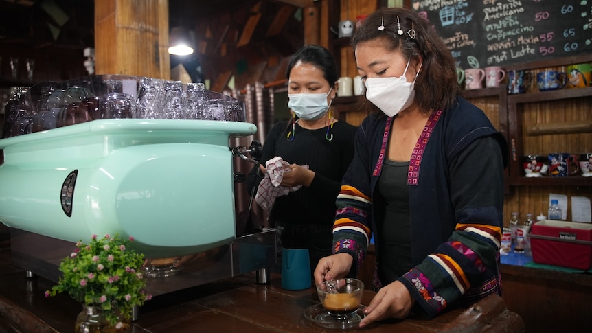 Two Thai women wearing masks stand behind a mint green industrial coffee machine in a cafe