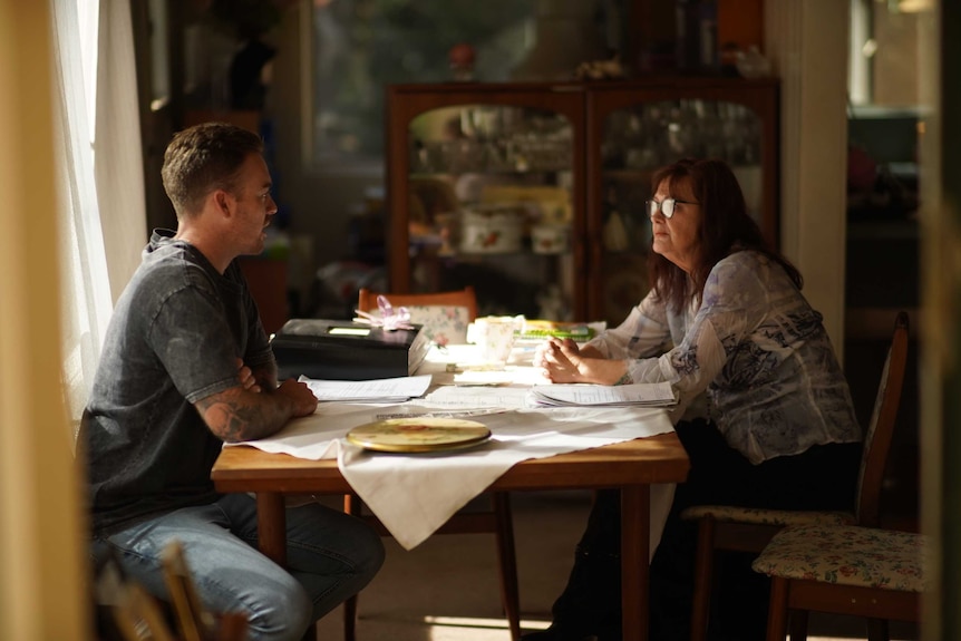 A man and a woman chat at a home dining table. the table is covered in paperwork, serious expressions on their face