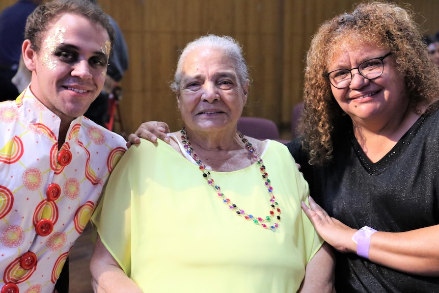 An elderly indigenous woman flanked by a younger indigenous woman and a young Indigenous man, all smiling