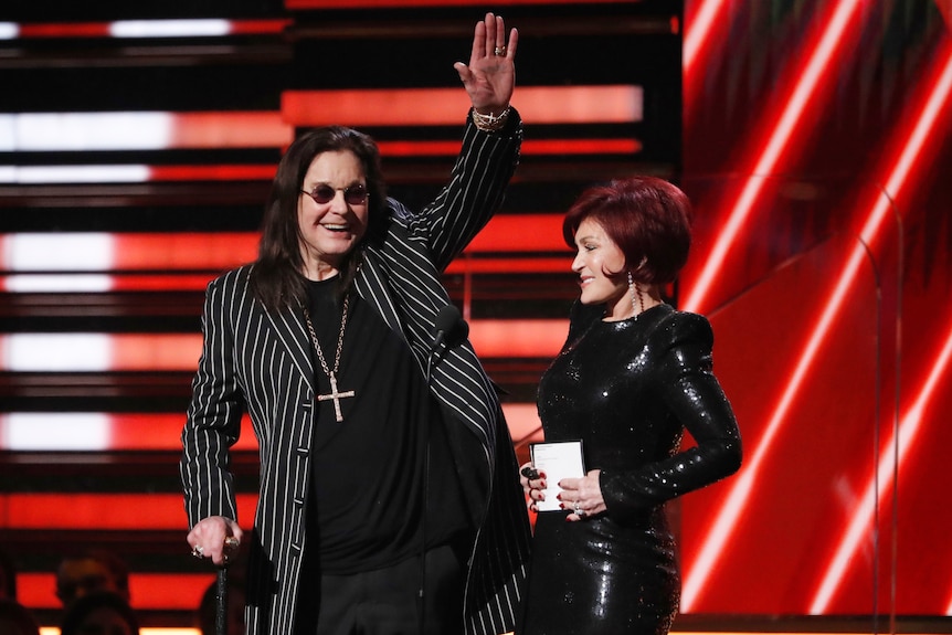 Ozzy Osbourne waves as his wife Sharon stands beside him on stage as they present an award at the Grammys.