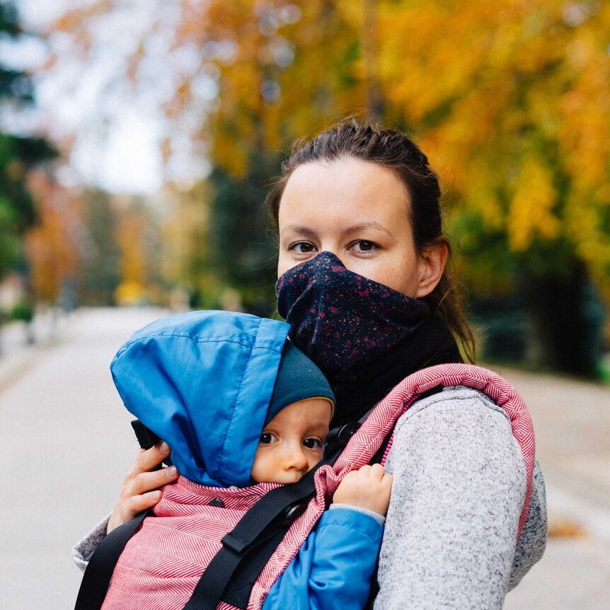 a woman holds a baby in a sling they both face the camera, the woman is wearing a face mask the baby looks apprehensive