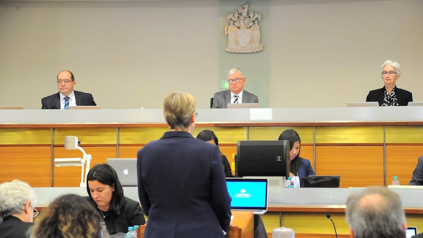 Three officials look over a court-type room. A lawyer can be seen, from behind, facing them.