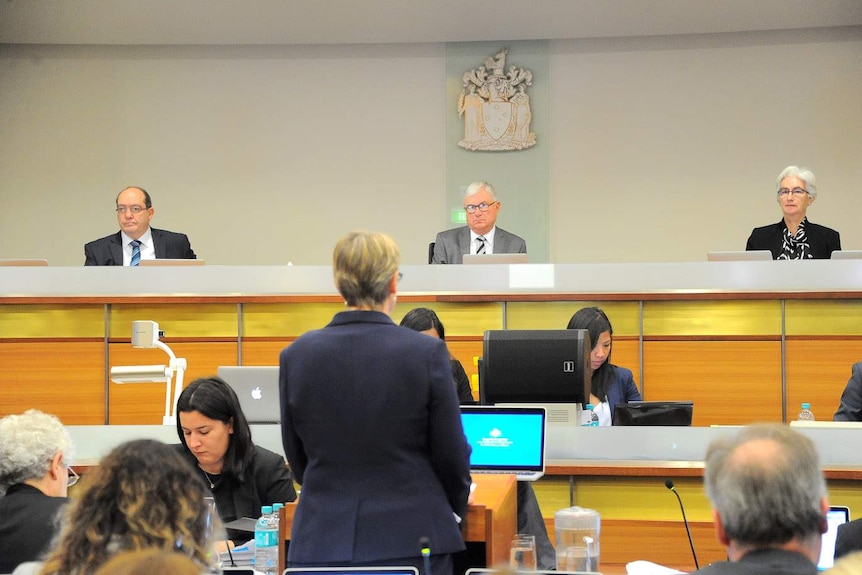 Three officials look over a court-type room. A lawyer can be seen, from behind, facing them.