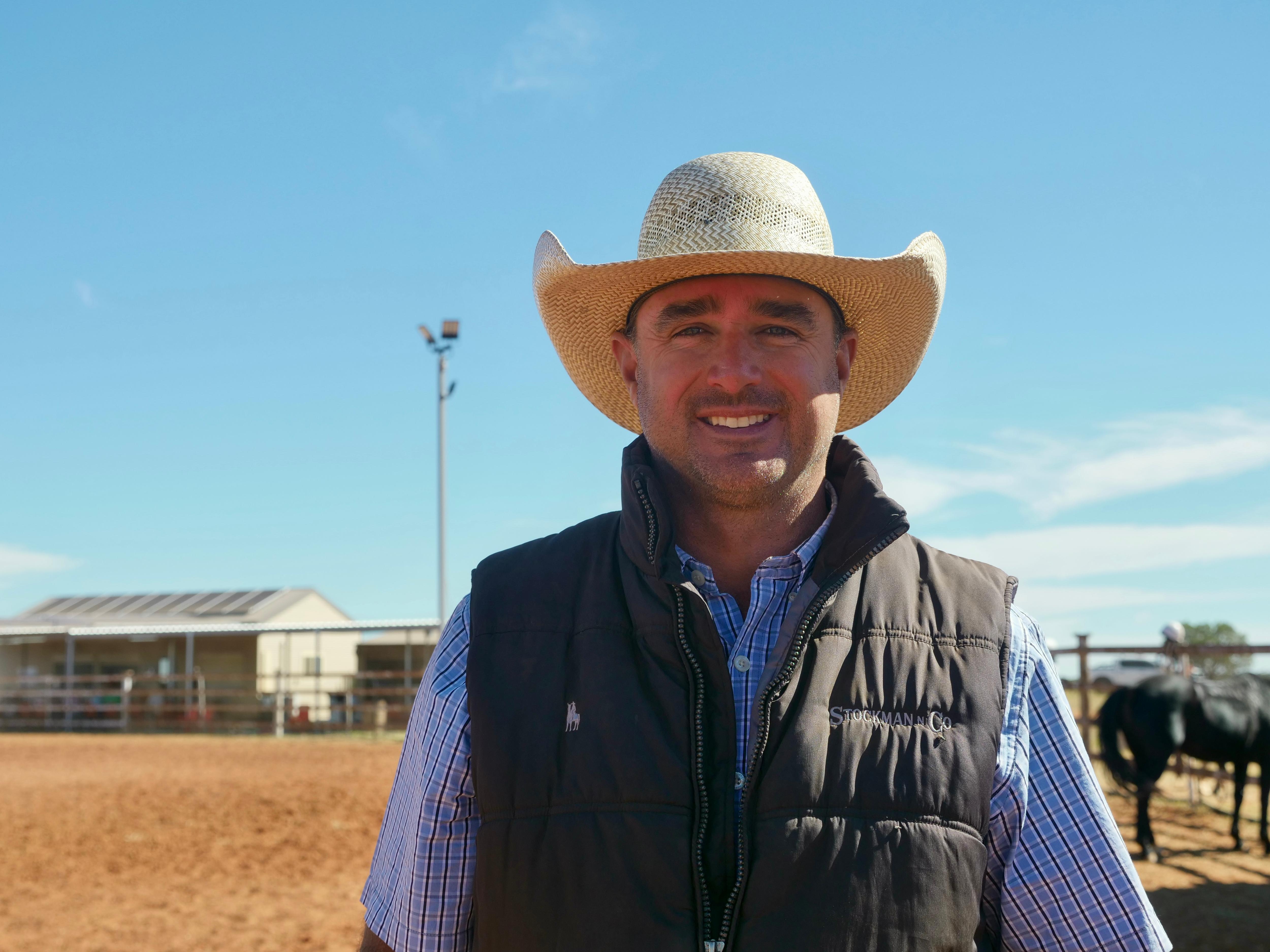 a man in a blue shirt with a black vest and hat on smiling at the camera with a red dirt background and blue skys