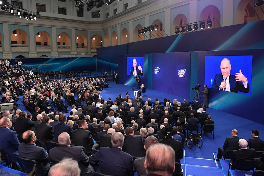 Russian President Vladimir Putin sits on stage and speaks to his supporters sitting in an arc around him.