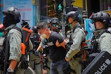 A man in a face mask grimaces with his hands held behind his back by police.