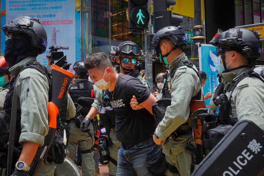 A man in a face mask grimaces with his hands held behind his back by police.