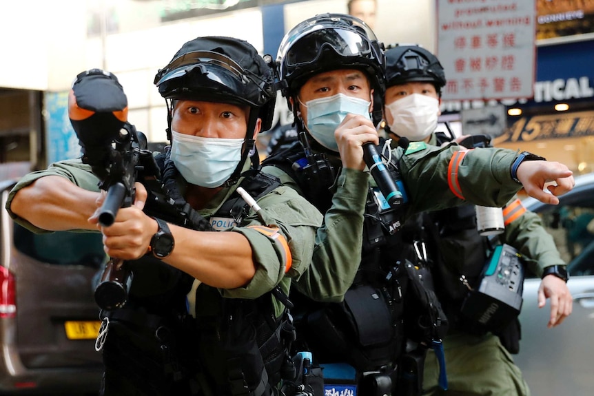 Hong Kong police in riot gear and masks, one carries a microphone while another raises a gun to fire balls of pepper spray.