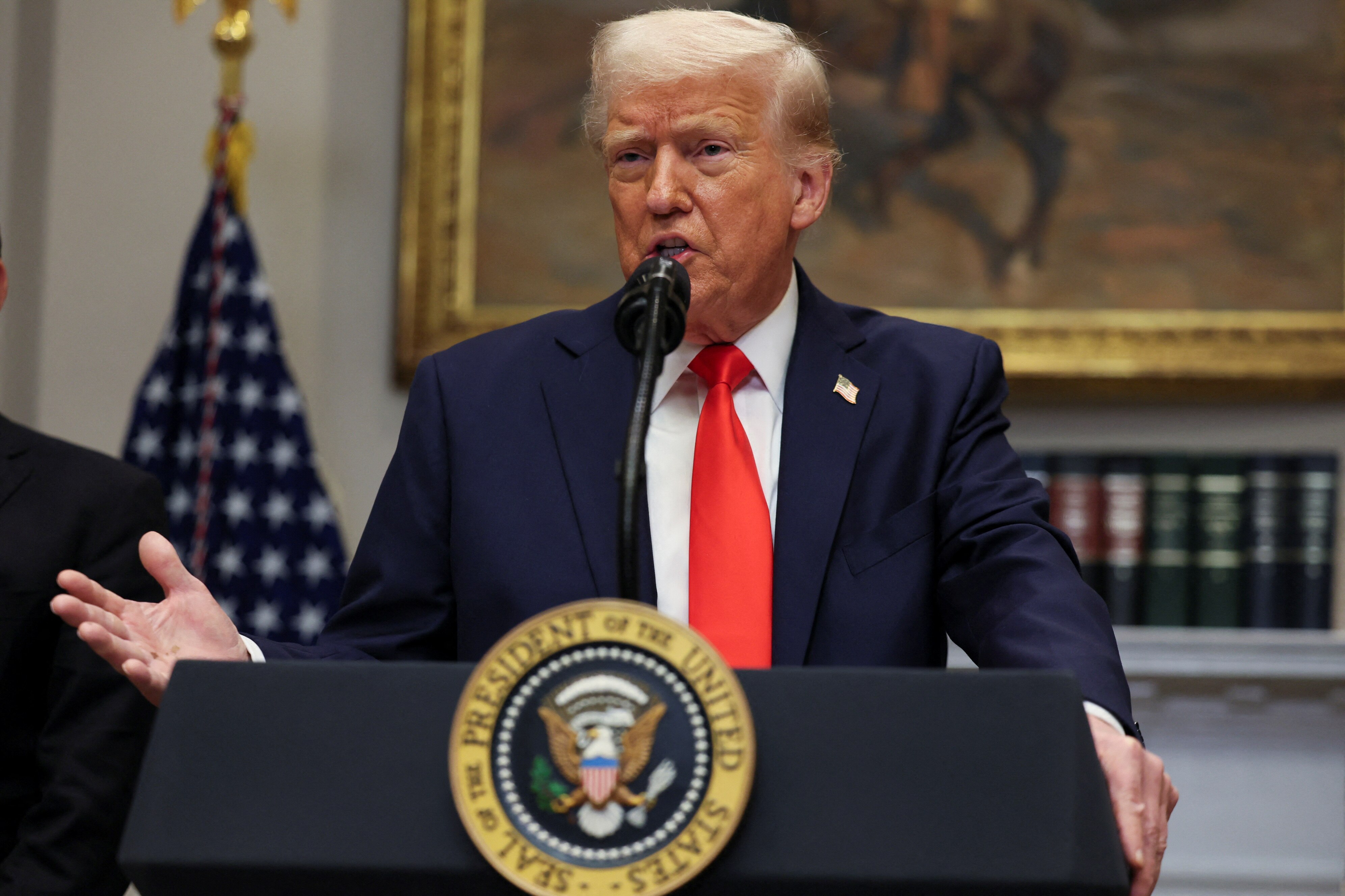 Donald Trump in a dark suit and red tie standing at a presidential lectern gesturing with his right hand in front of a US flag